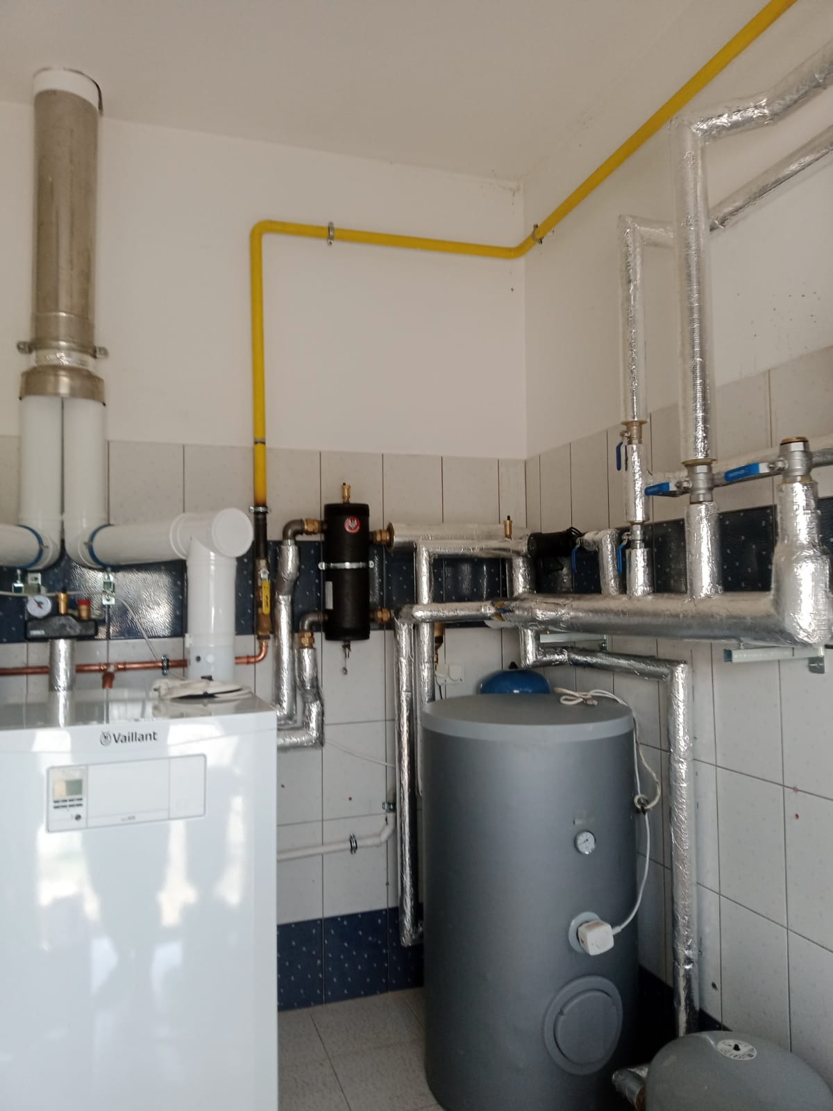 Biały piec grzewczy Vaillant z szarym zasobnikiem wody, rurami w izolacji i żółtą instalacją gazową w pomieszczeniu wyłożonym częściowo kafelkami.