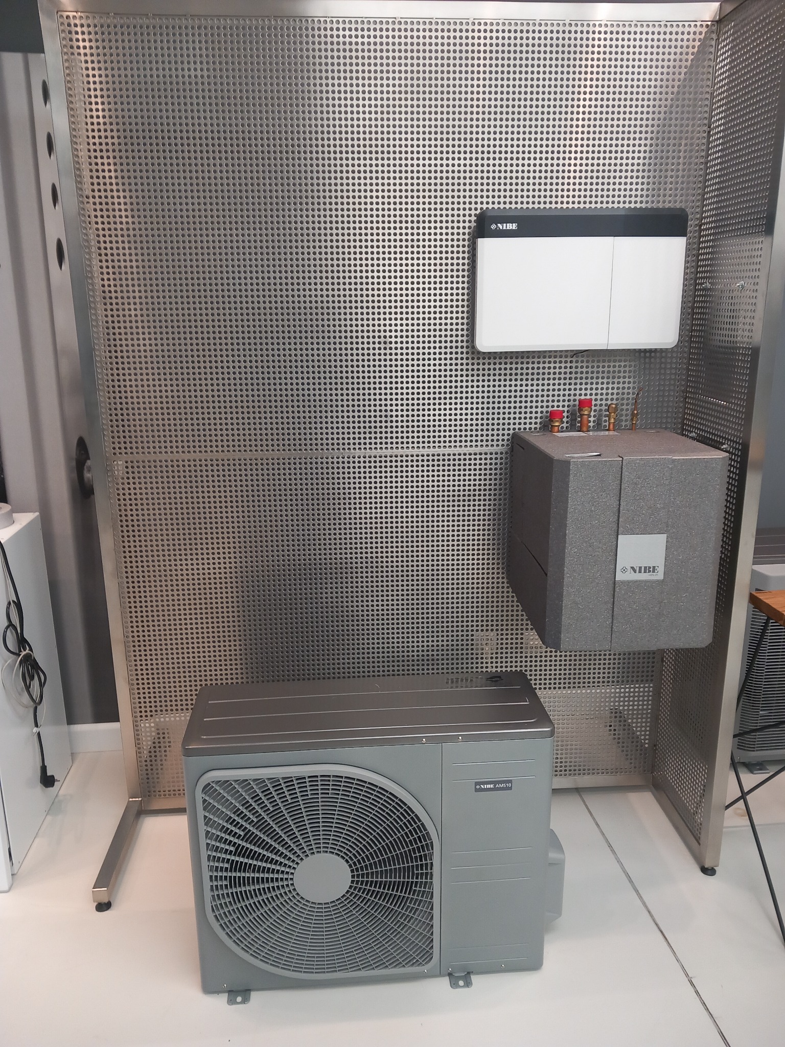 Szara pompa ciepła Nibe AMS10, składająca się z jednostki zewnętrznej z wentylatorem, modułu wewnętrznego z rurami i zasobnika, umieszczona na tle perforowanej metalowej ścianki ekspozycyjnej.