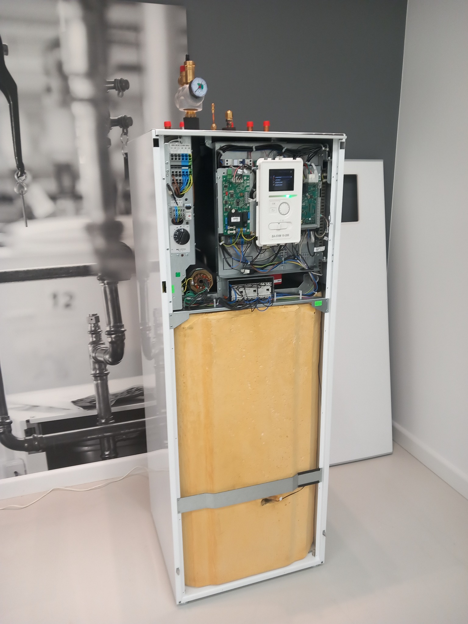 Otwarta obudowa urządzenia grzewczego z widocznymi podzespołami elektronicznymi, izolacją termiczną i elementami instalacji, na tle zdjęcia instalacji hydraulicznej.