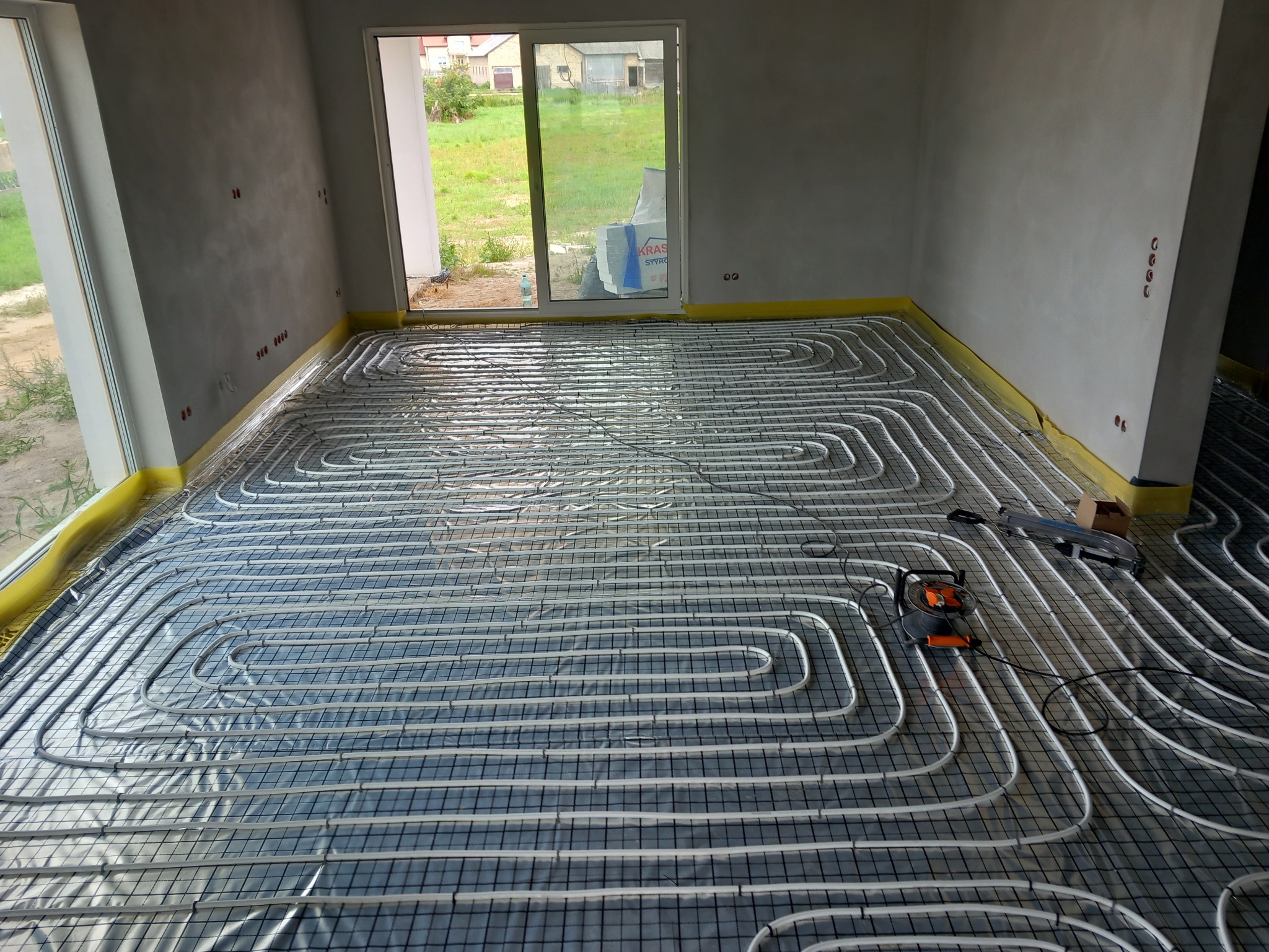 Rozłożone rury ogrzewania podłogowego na siatce w surowym wnętrzu z widokiem na zewnątrz przez okno balkonowe, widoczny rozwijak do rur.