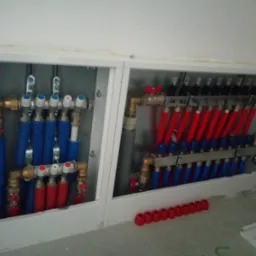 Rozdzielacz ogrzewania podłogowego w zabudowie z rurami w kolorze czerwonym i niebieskim, widoczne zawory i złączki