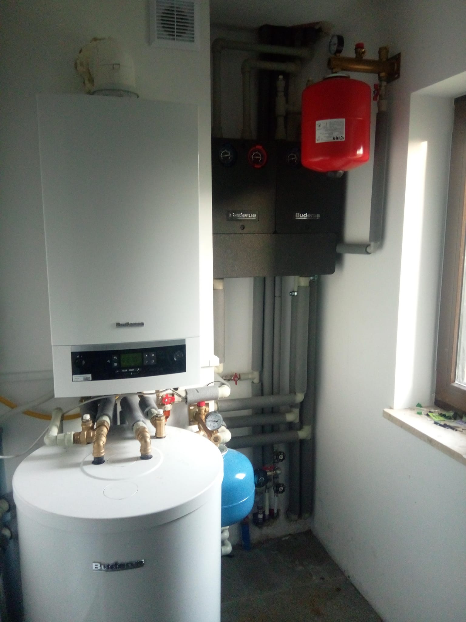 Kompaktowa kotłownia z białym kotłem gazowym kondensacyjnym Buderus, szarym modułem sterującym i czerwonym zbiornikiem wyrównawczym, instalacja z szarymi rurami w izolacji, widok z boku.