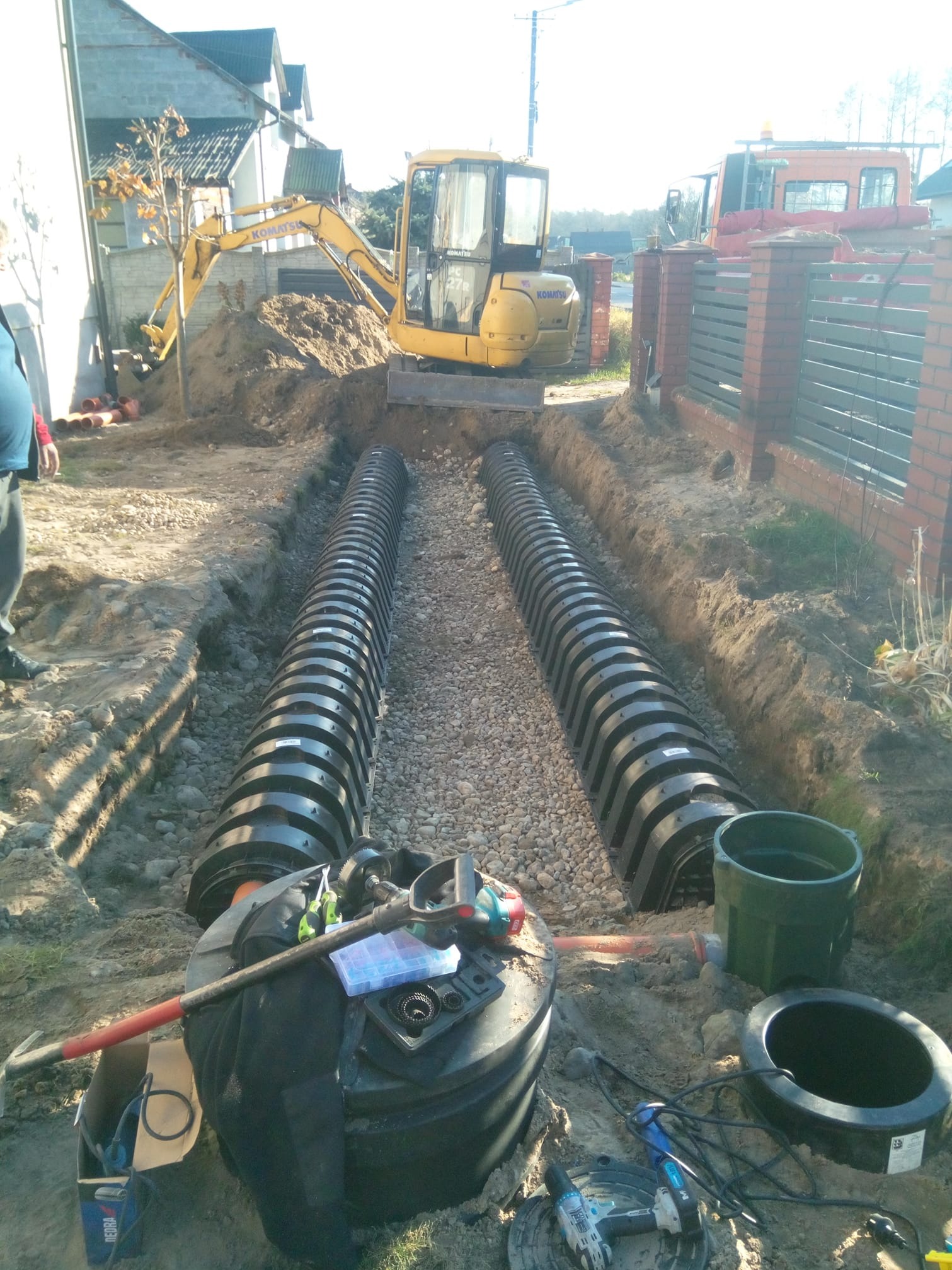 Dwa rzędy czarnych rur drenażowych ułożone w wykopie wypełnionym żwirem, z żółtą koparką Komatsu w tle i narzędziami na pokrywie zbiornika z tworzywa sztucznego na pierwszym planie.