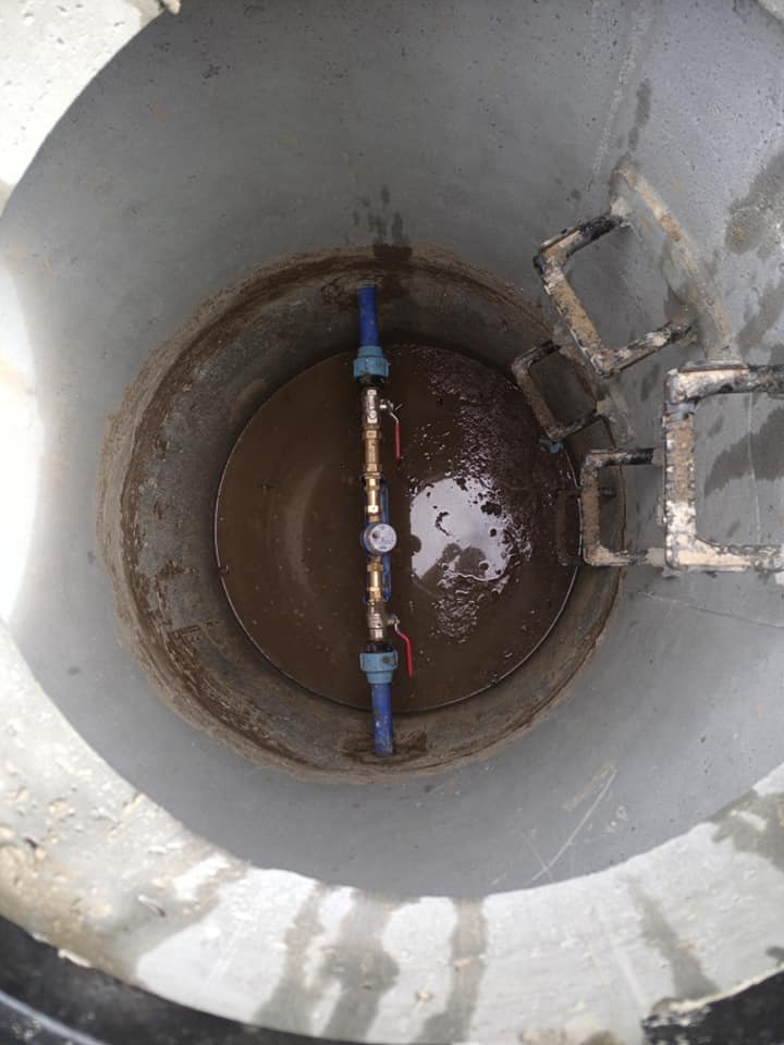 Wnętrze betonowego kręgu studni z instalacją wodną: niebieskie rury, zawory z czerwonymi uchwytami i manometr, częściowo zanurzone w brudnej wodzie, drabinka z metalowych prętów.