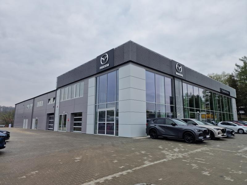 Nowoczesny salon samochodowy Mazda z szarymi panelami i dużymi oknami, zaparkowane auta na pierwszym planie, pochmurne niebo w tle. Widok z zewnątrz.