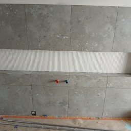 Rem-Pat - Ściana łazienki wyłożona szarymi płytkami imitującymi kamień, z wnęką wykończoną białymi, ryflowanymi panelami. Widoczne wystające rurki instalacji wodnej w kolorze czerwonym i niebieskim.