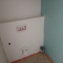 Rem-Pat - Stelaż podtynkowy WC zamontowany na białej ściance działowej z płyty kartonowo-gipsowej, widoczne przyłącza i fragment niebieskiej ściany z płyt gipsowych w surowym stanie.