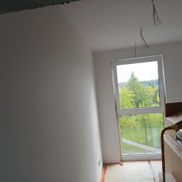 Rem-Pat - Świeżo pomalowane na biało ściany i sufit w pokoju z dużym oknem, przez które widać zieleń drzew; widoczne wystające kable ze stropu i zabezpieczona podłoga.