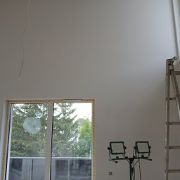 Rem-Pat - Przygotowane do malowania wnętrze z oknem zabezpieczonym taśmą malarską, oświetlone halogenami budowlanymi, z drabiną opartą o ścianę.