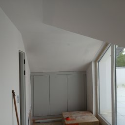 Rem-Pat - Wnętrze poddasza po remoncie, widoczne białe ściany, zabudowa szafy wnękowej z szarymi frontami, duże okno z widokiem na zewnątrz i kartonowe pudło z pomarańczową taśmą na podłodze.