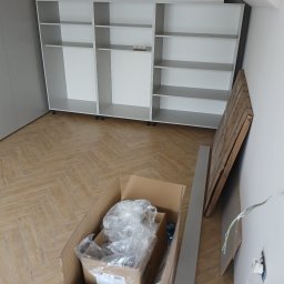 Rem-Pat - Montaż szafy wnękowej z szarymi frontami i białymi półkami, widoczna kartonowa podłoga, elementy wykończeniowe i wystające kable elektryczne.