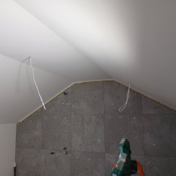 Rem-Pat - W trakcie układania szarych płytek na ścianie w łazience ze skosem w Toruniu, widoczne kable wystające ze ściany i taśma zabezpieczająca.