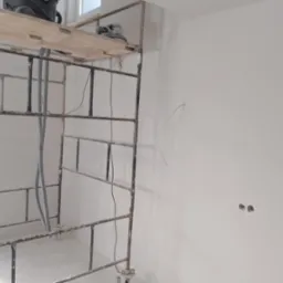 Metalowe rusztowanie z szlifierką na górze, przewody i otwory w ścianie sugerują przygotowanie do prac wykończeniowych w pomieszczeniu.