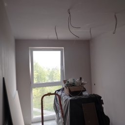 Rem-Pat - Pokój w trakcie prac wykończeniowych: świeżo pomalowane ściany na jasny kolor, wystające kable z sufitu, okno z widokiem na zieleń, drabina i materiały budowlane przykryte folią.