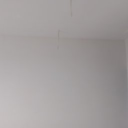 Rem-Pat - Świeżo pomalowana na biało ściana w rogu pokoju z widocznymi kablami zwisającymi z sufitu i surowym wykończeniem ościeżnicy drzwiowej.