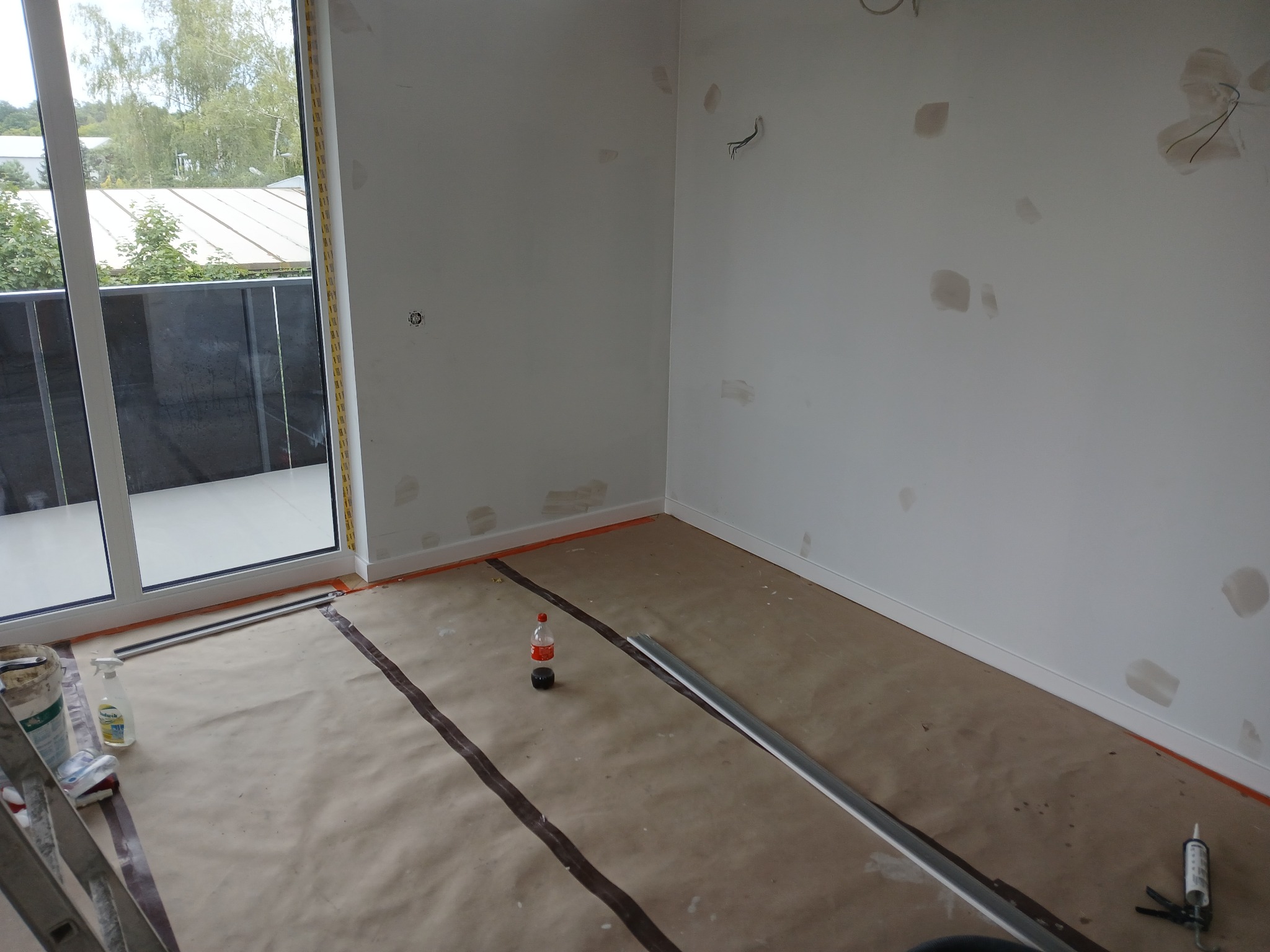 Pokój w trakcie przygotowań do prac wykończeniowych, ściany z widocznymi miejscami po szpachlowaniu, podłoga zabezpieczona papierem, widoczna butelka i narzędzia.