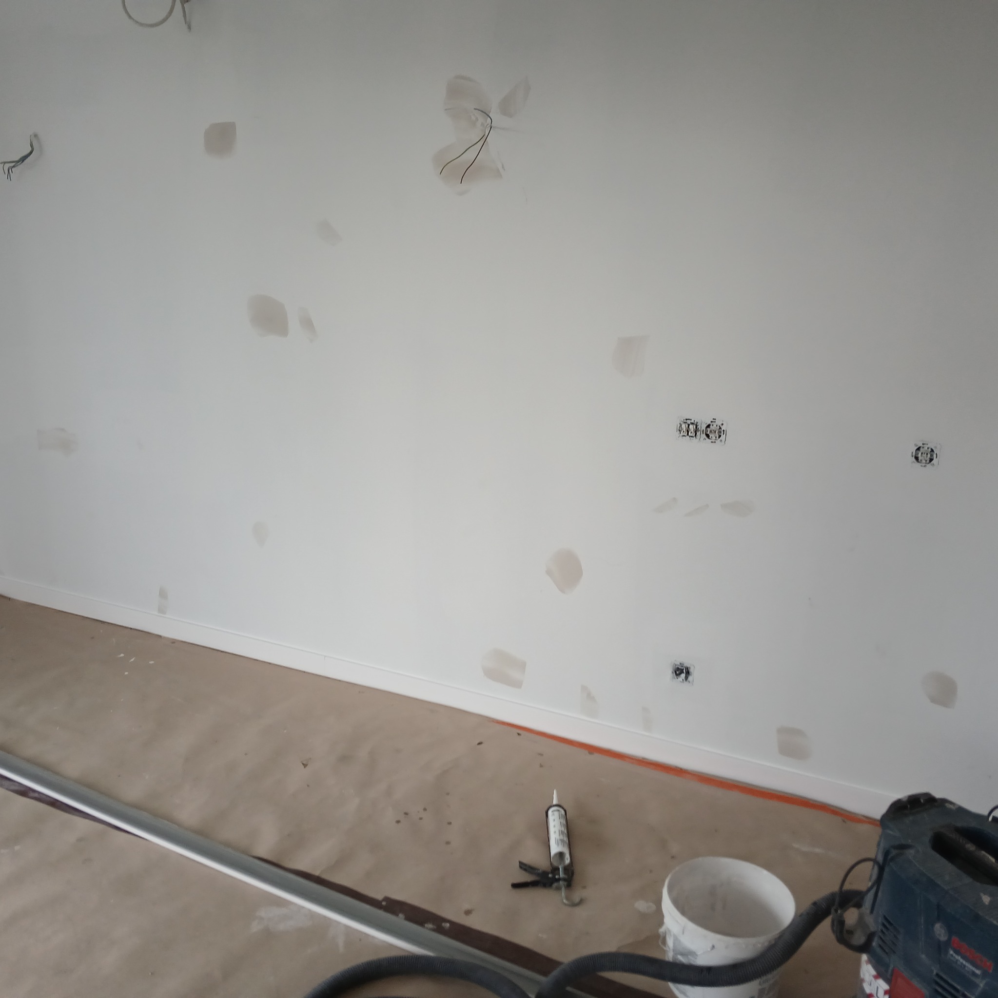 Biała ściana z widocznymi miejscami po szpachlowaniu, przygotowana do dalszych prac. Na podłodze zabezpieczającej leży listwa przypodłogowa, tuba z silikonem oraz wiadro i odkurzacz przemysłowy.