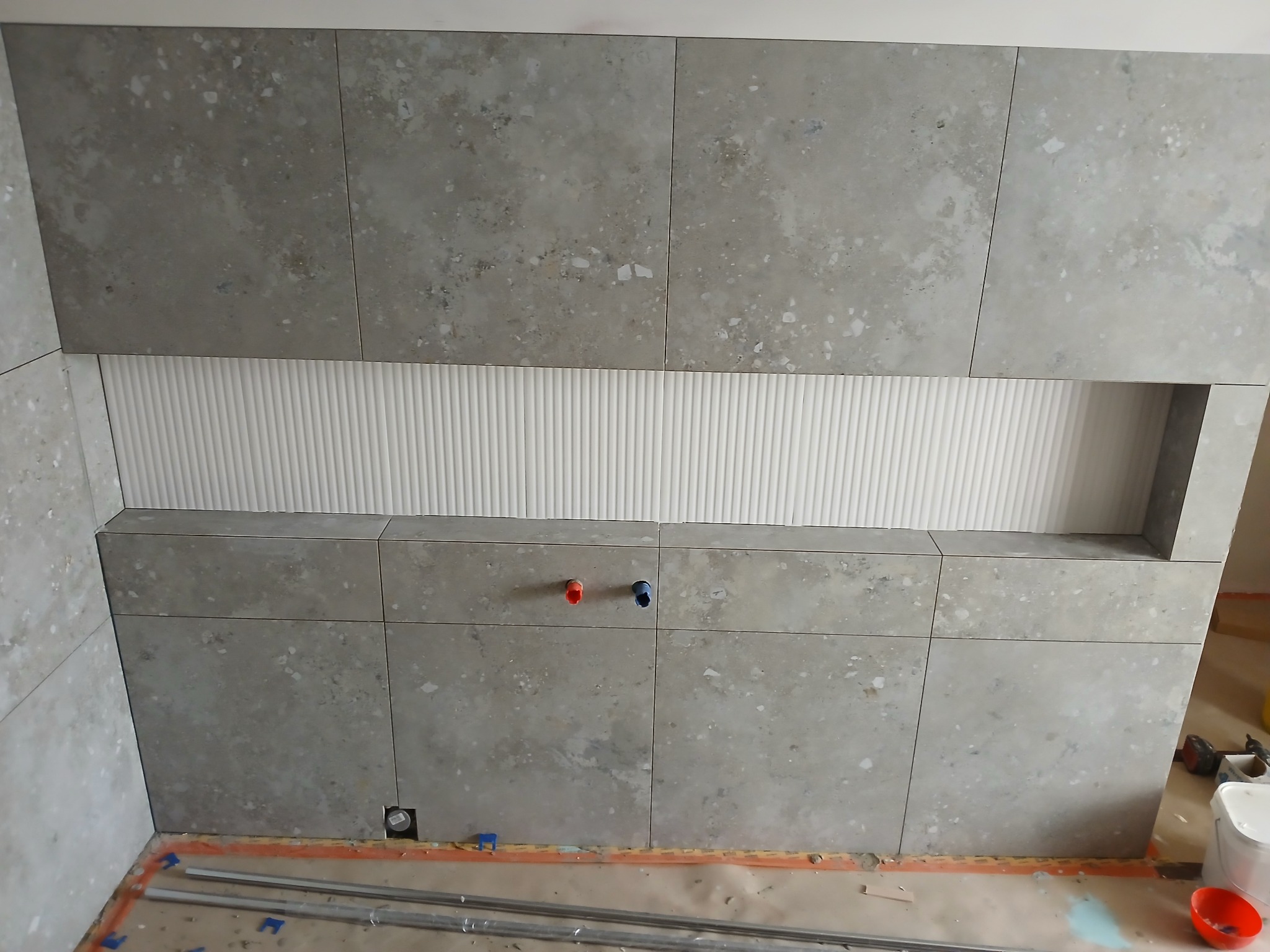 Ściana łazienki wyłożona szarymi płytkami imitującymi kamień, z wnęką wykończoną białymi, ryflowanymi panelami. Widoczne wystające rurki instalacji wodnej w kolorze czerwonym i niebieskim.