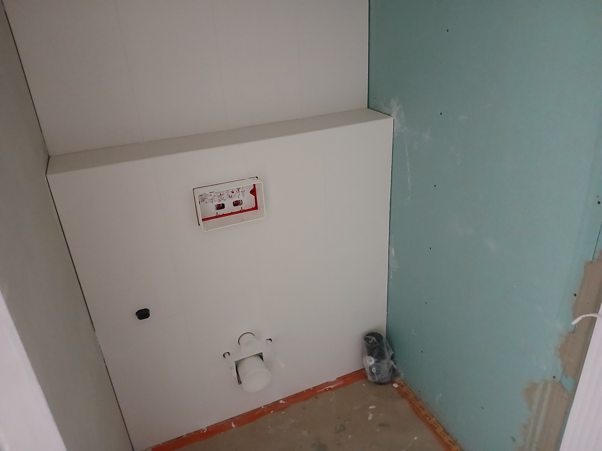Stelaż podtynkowy WC zamontowany na białej ściance działowej z płyty kartonowo-gipsowej, widoczne przyłącza i fragment niebieskiej ściany z płyt gipsowych w surowym stanie.