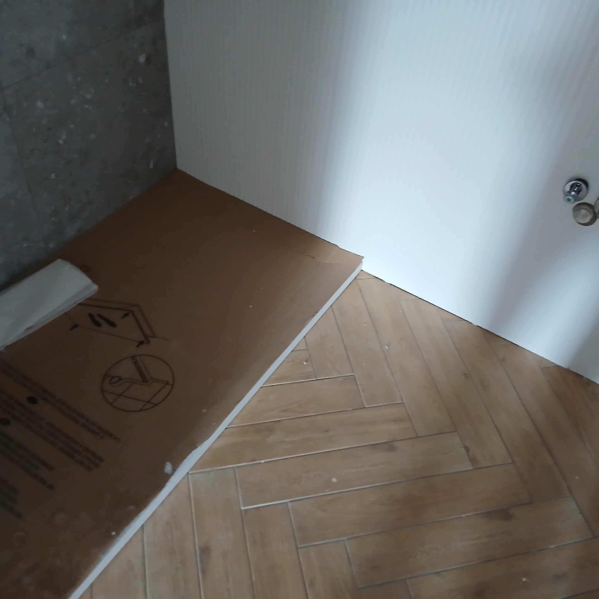 Karton z płytami gipsowo-kartonowymi i fragmentem ułożonej podłogi z płytek imitujących drewno w jodełkę, widok z góry w rogu pomieszczenia.