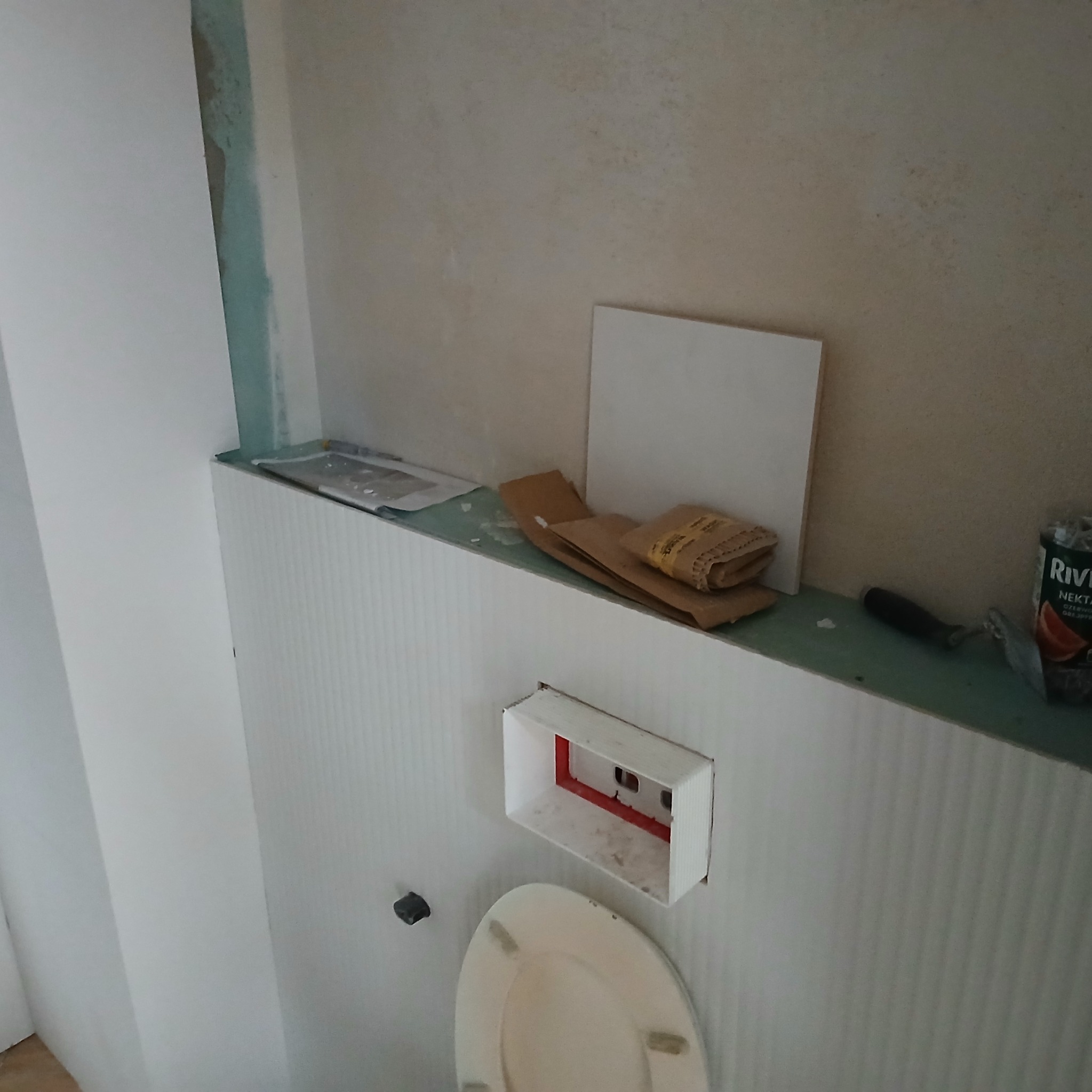 Niedokończona łazienka z widoczną instalacją podtynkową WC, płytką, szpachelką i materiałami ściernymi na nierównym podłożu.