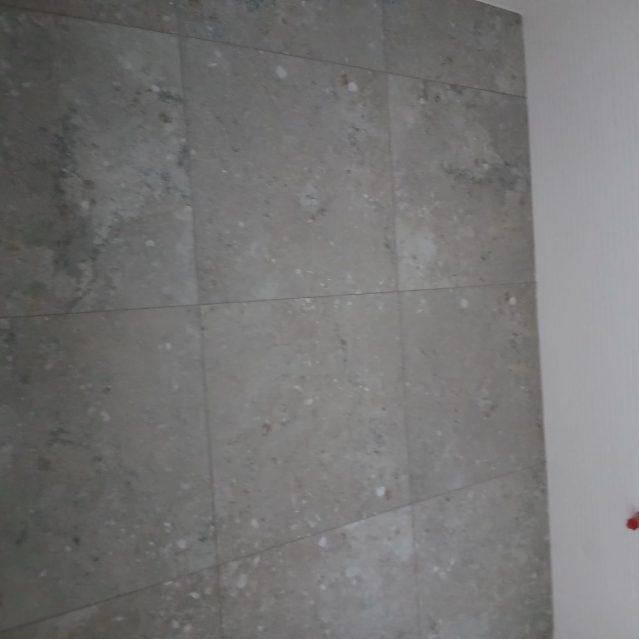 Ściana wyłożona szarymi, marmurkowymi płytkami ceramicznymi z widocznymi fugami, przylegająca do białej, gładkiej ściany z elementem instalacji elektrycznej.