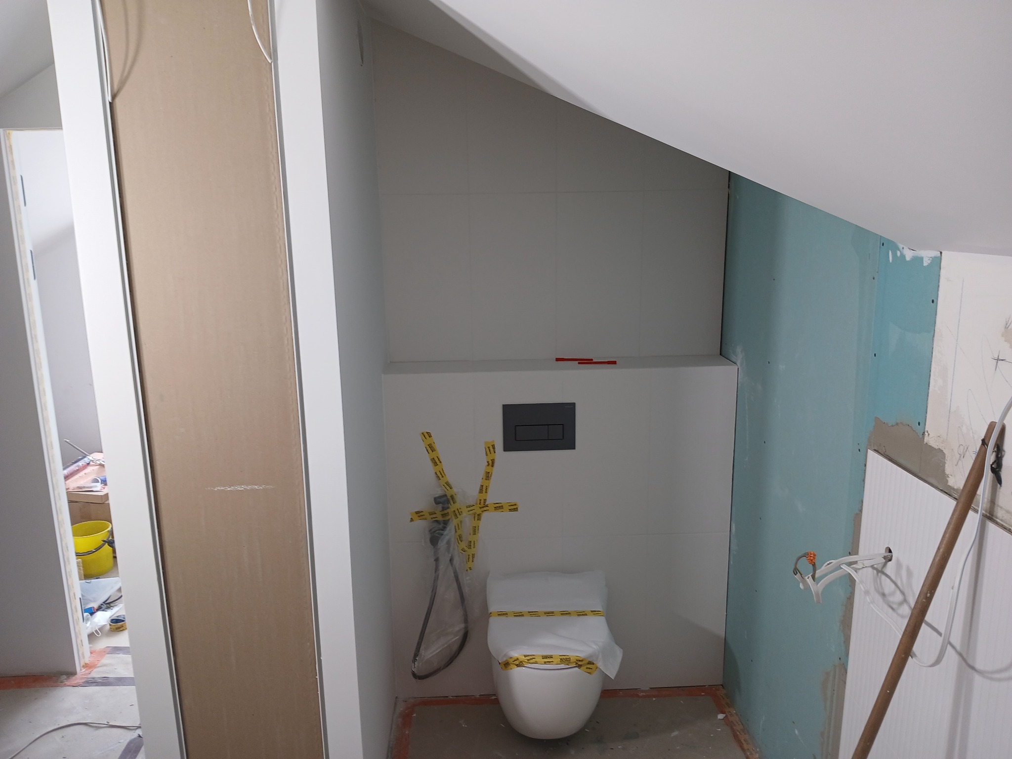 Łazienka w trakcie prac: biała toaleta zabezpieczona taśmą, szara spłuczka podtynkowa, białe płytki na ścianie, fragment ściany z płyt kartonowo-gipsowych w kolorze niebieskim, widoczne przewody...