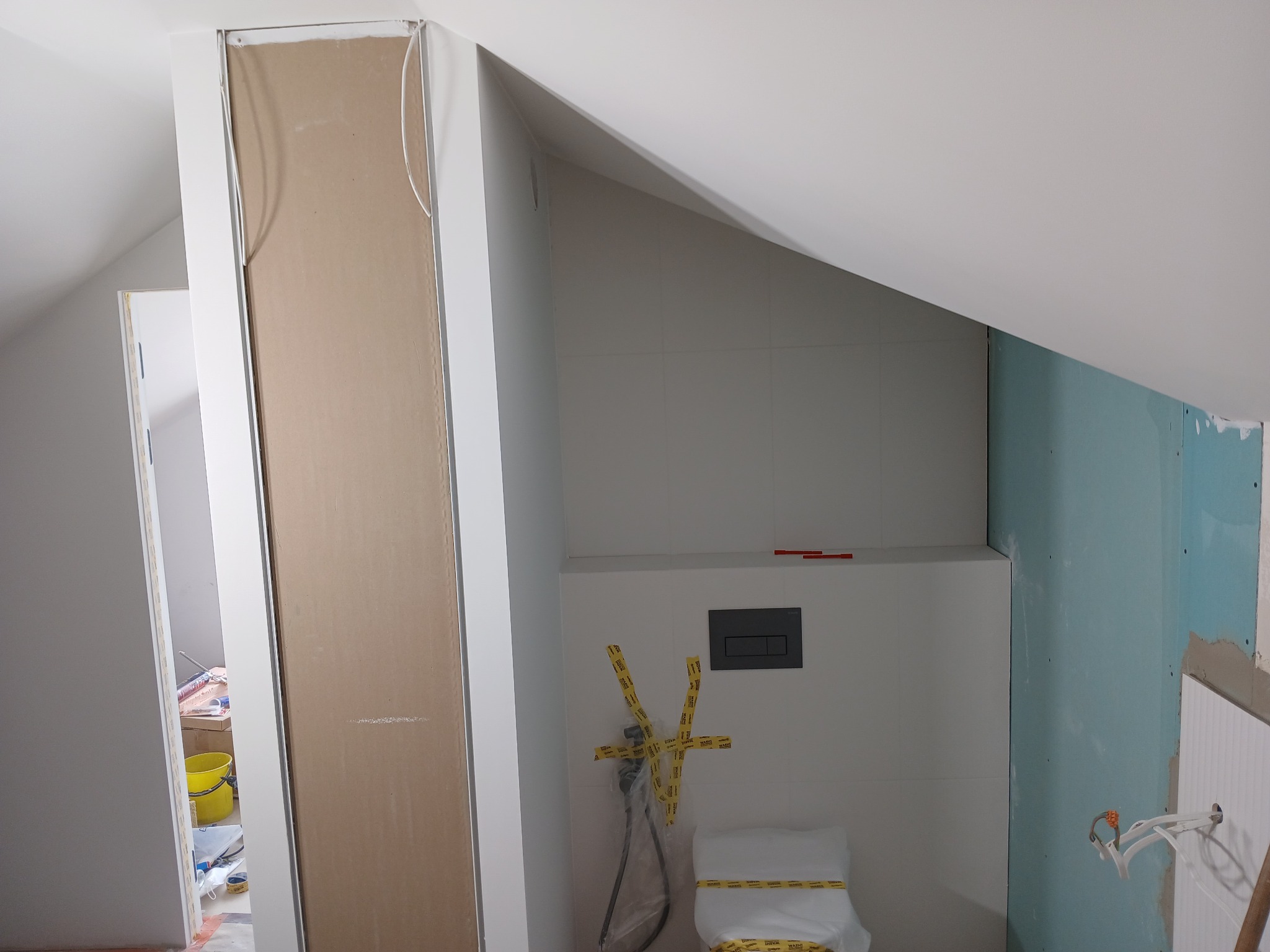 Łazienka w trakcie prac: obudowa z płyty karton gips, częściowo wykończone płytki, zabezpieczona taśmą toaleta, widoczne instalacje.