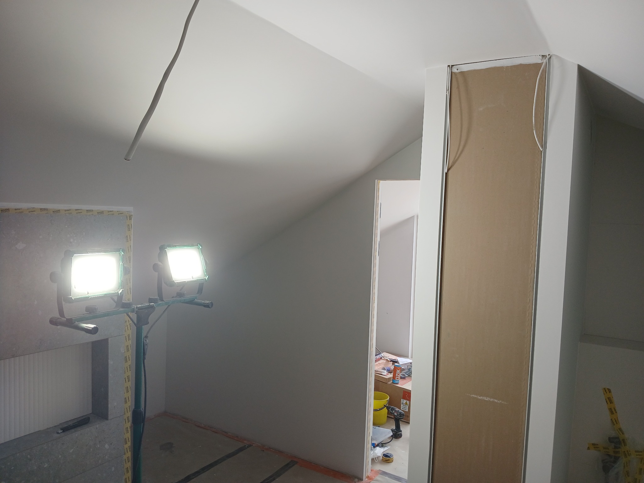 Podświetlone halogenami wnętrze poddasza w trakcie prac wykończeniowych, widoczne płyty kartonowo-gipsowe, narzędzia i materiały budowlane, pomarańczowa taśma wyznaczająca obszar roboczy na podłodze.