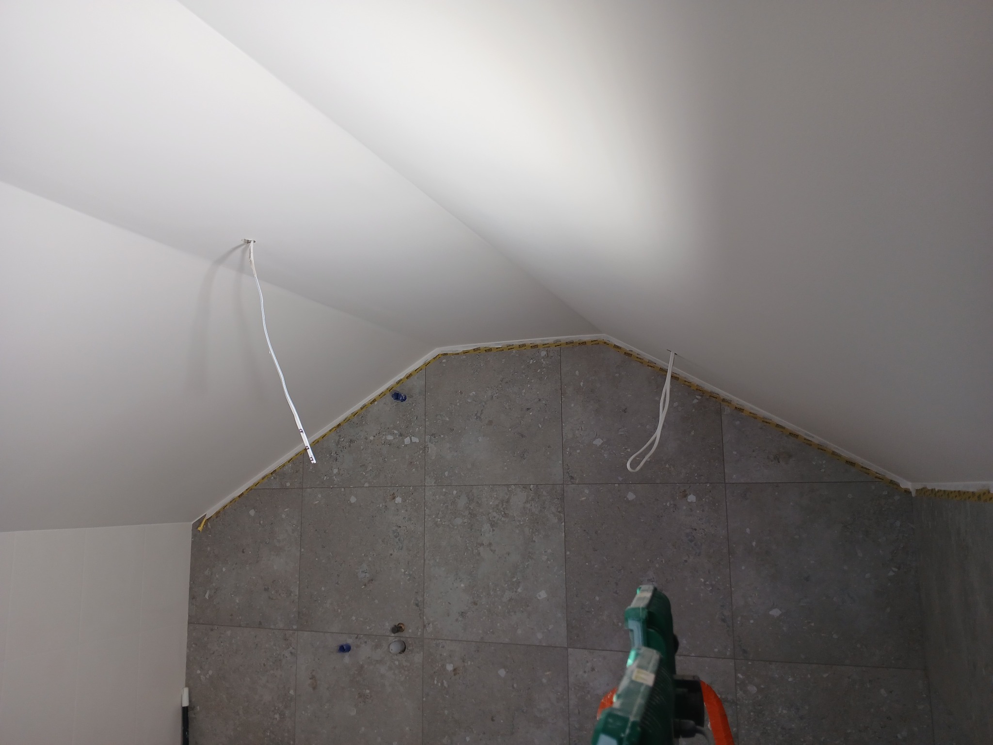 W trakcie układania szarych płytek na ścianie w łazience ze skosem w Toruniu, widoczne kable wystające ze ściany i taśma zabezpieczająca.