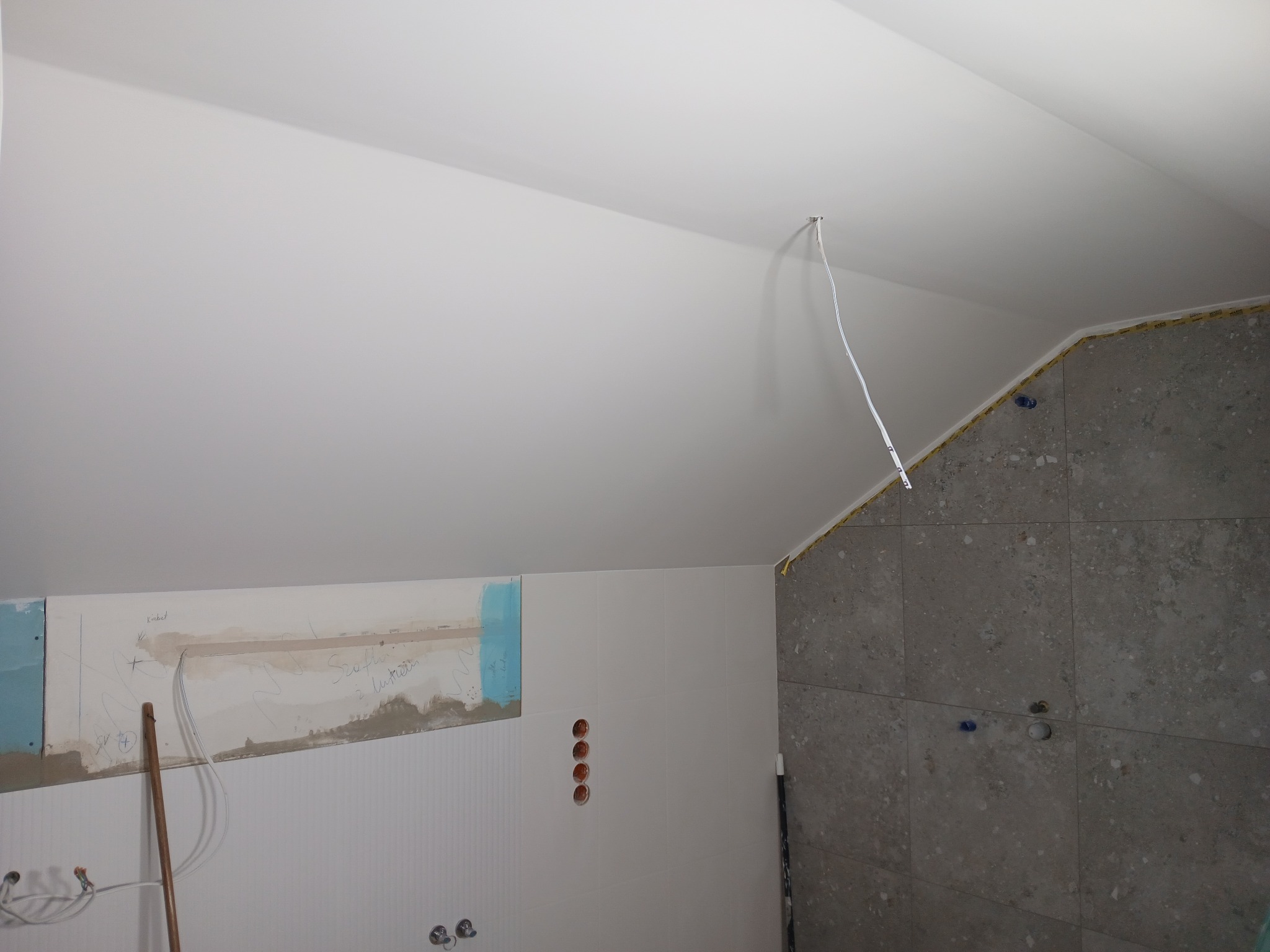 Remont łazienki w trakcie: częściowo położone szare płytki na ścianie, białe płytki z pionowymi żłobieniami, fragment ściany z nieotynkowanym obszarem z odręcznymi notatkami i widocznymi przewodami...