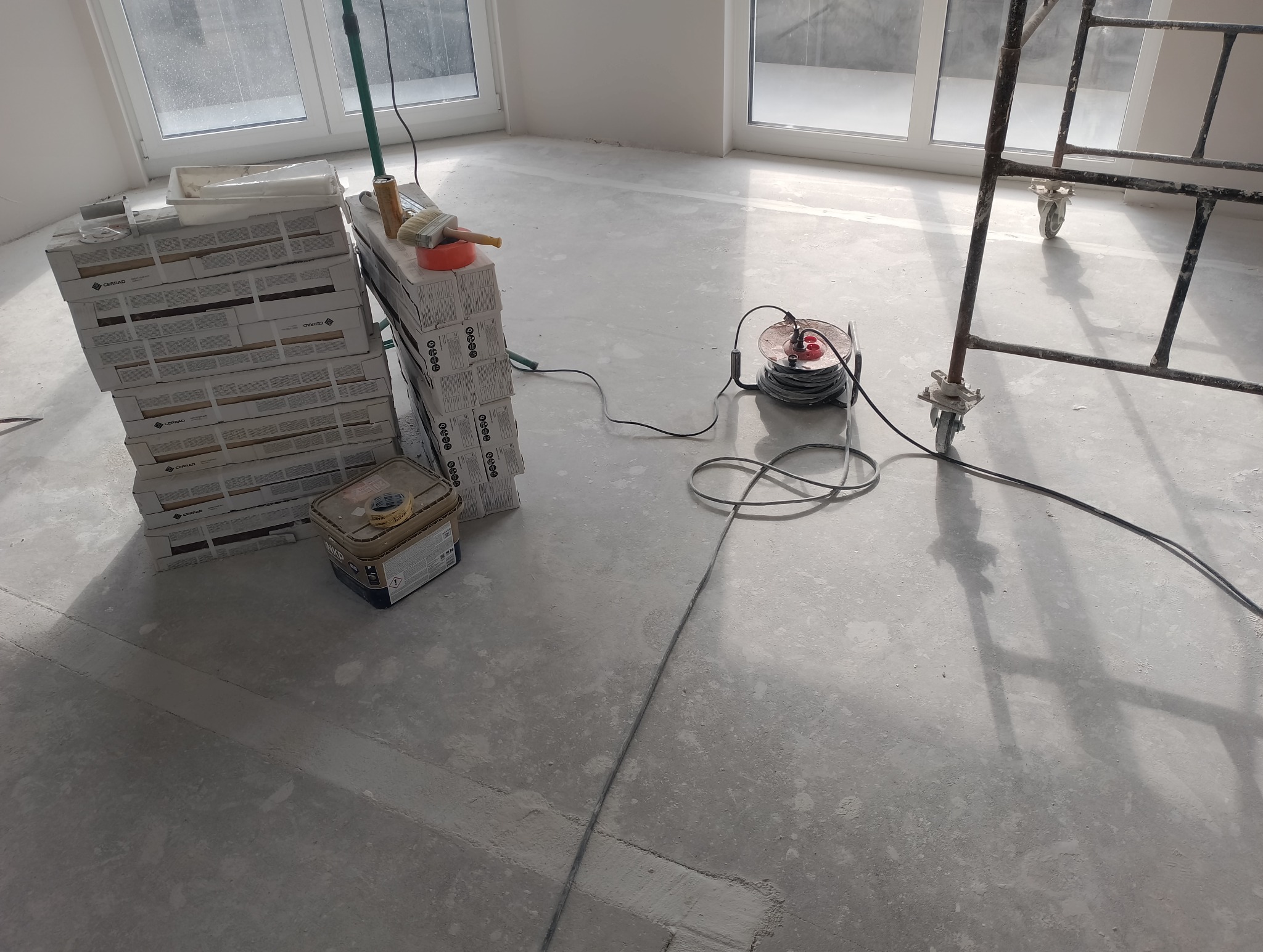 Przygotowany do położenia gresu szary, betonowy podkład podłogowy w jasnym pomieszczeniu z dwoma oknami, obok stos paczek płytek Cerrad, puszka z klejem, przedłużacz i rusztowanie.