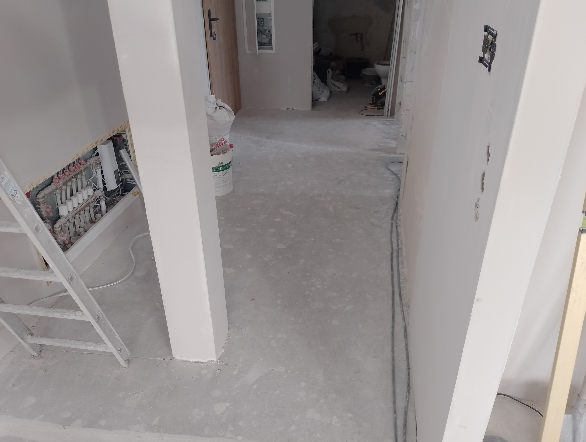 Wnętrze w trakcie remontu: widoczne ściany po szpachlowaniu, przygotowane do malowania, instalacja elektryczna w ścianie, drabina malarska i worki z zaprawą w tle.