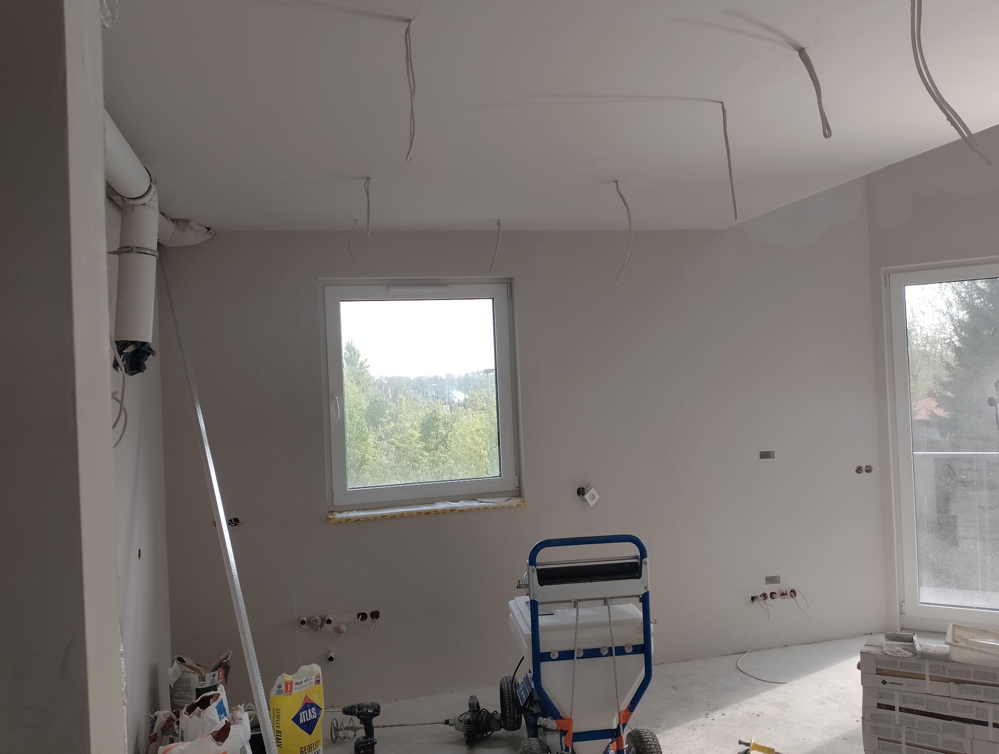 Wnętrze w trakcie prac wykończeniowych: ściany przygotowane do malowania, widoczna maszyna do nakładania gładzi, okno z widokiem na zieleń, wystające przewody elektryczne i worki z materiałami...