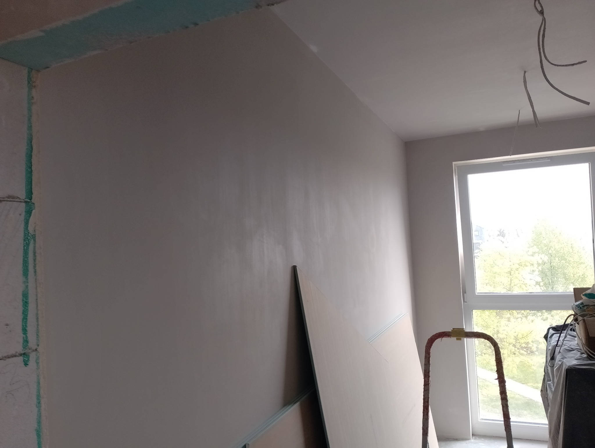 Gładka, wyszpachlowana ściana w jasnym odcieniu szarości w trakcie prac wykończeniowych, z widocznymi przewodami elektrycznymi wystającymi z sufitu i fragmentem płyty kartonowo-gipsowej opartej...