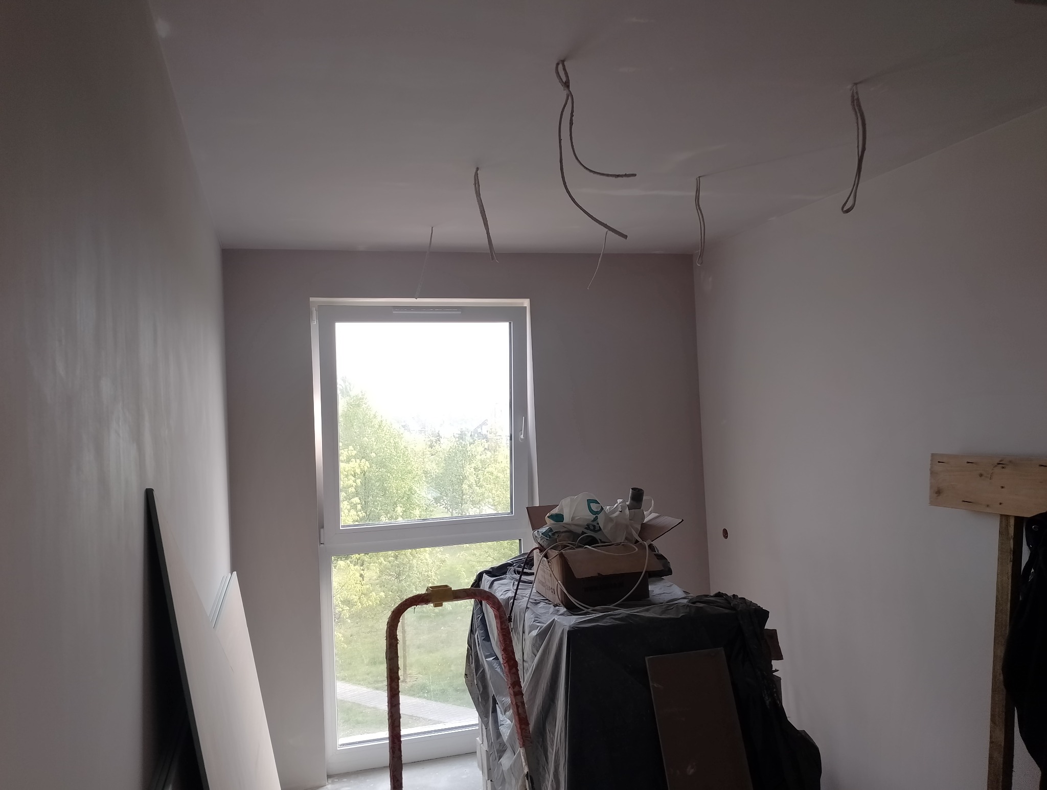 Pokój w trakcie prac wykończeniowych: świeżo pomalowane ściany na jasny kolor, wystające kable z sufitu, okno z widokiem na zieleń, drabina i materiały budowlane przykryte folią.