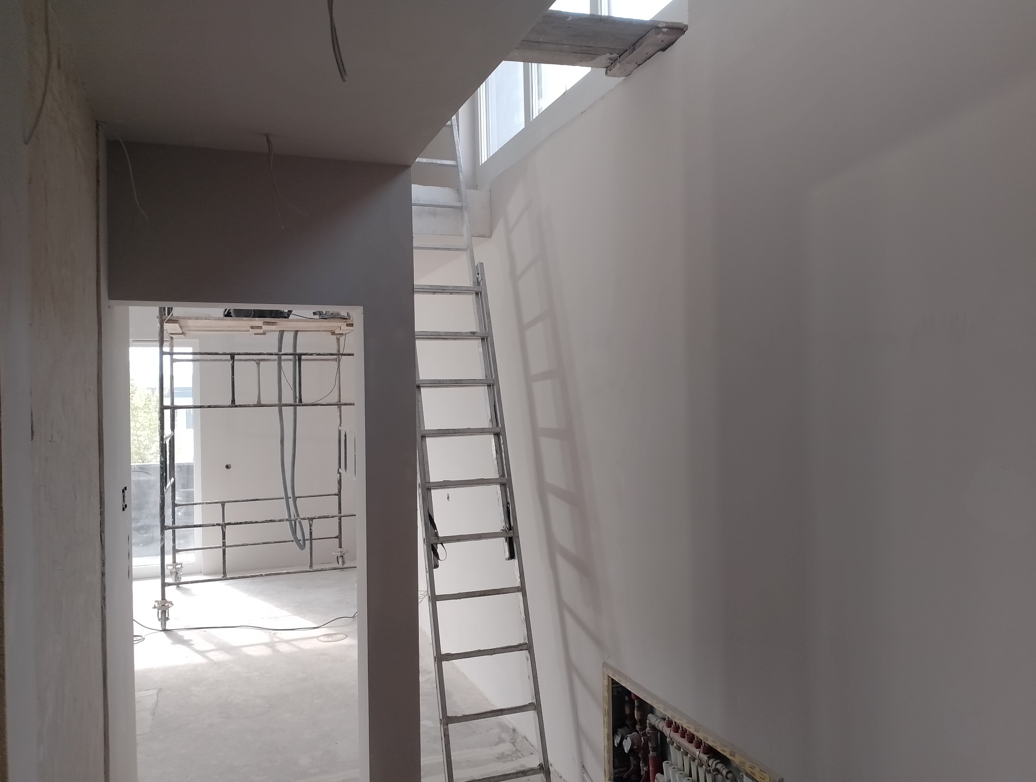 Wnętrze w trakcie remontu: białe ściany po szpachlowaniu, aluminiowa drabina oparta o ścianę, rusztowanie w sąsiednim pomieszczeniu, instalacja grzewcza w ścianie