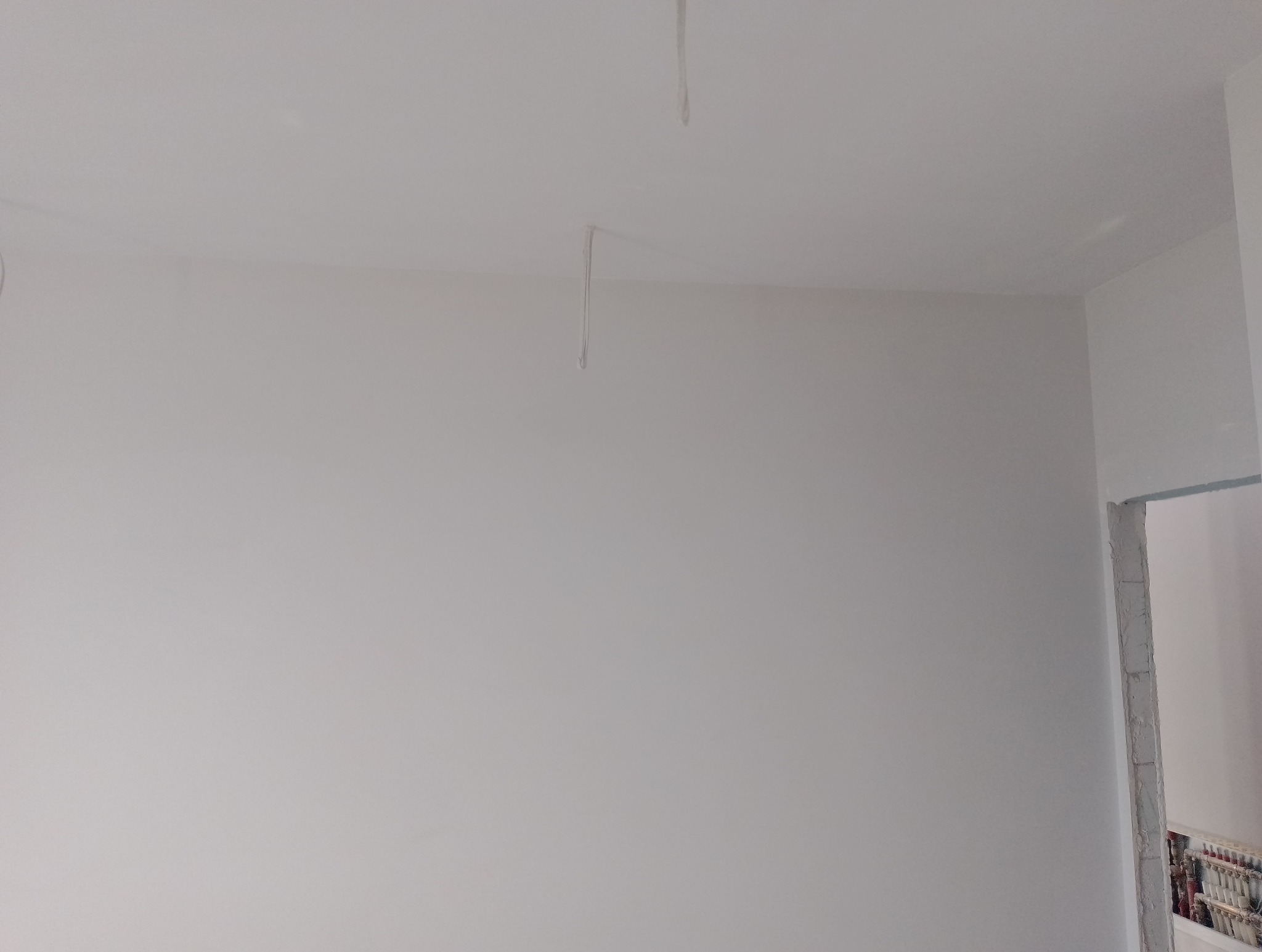 Świeżo pomalowana na biało ściana w rogu pokoju z widocznymi kablami zwisającymi z sufitu i surowym wykończeniem ościeżnicy drzwiowej.