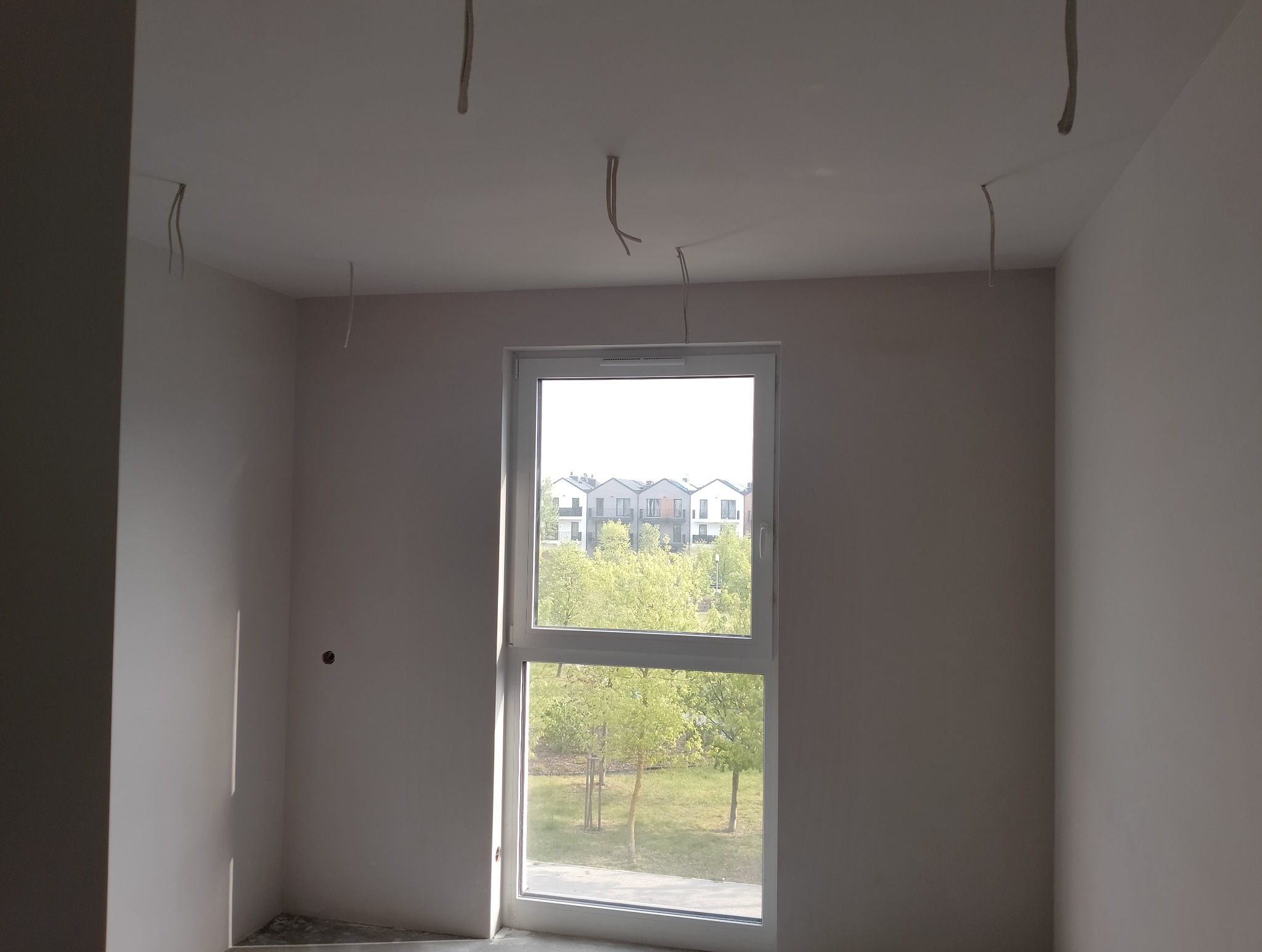 Pokój w stanie deweloperskim z białymi ścianami, widoczne wystające kable z sufitu, okno z widokiem na osiedle i zieleń.