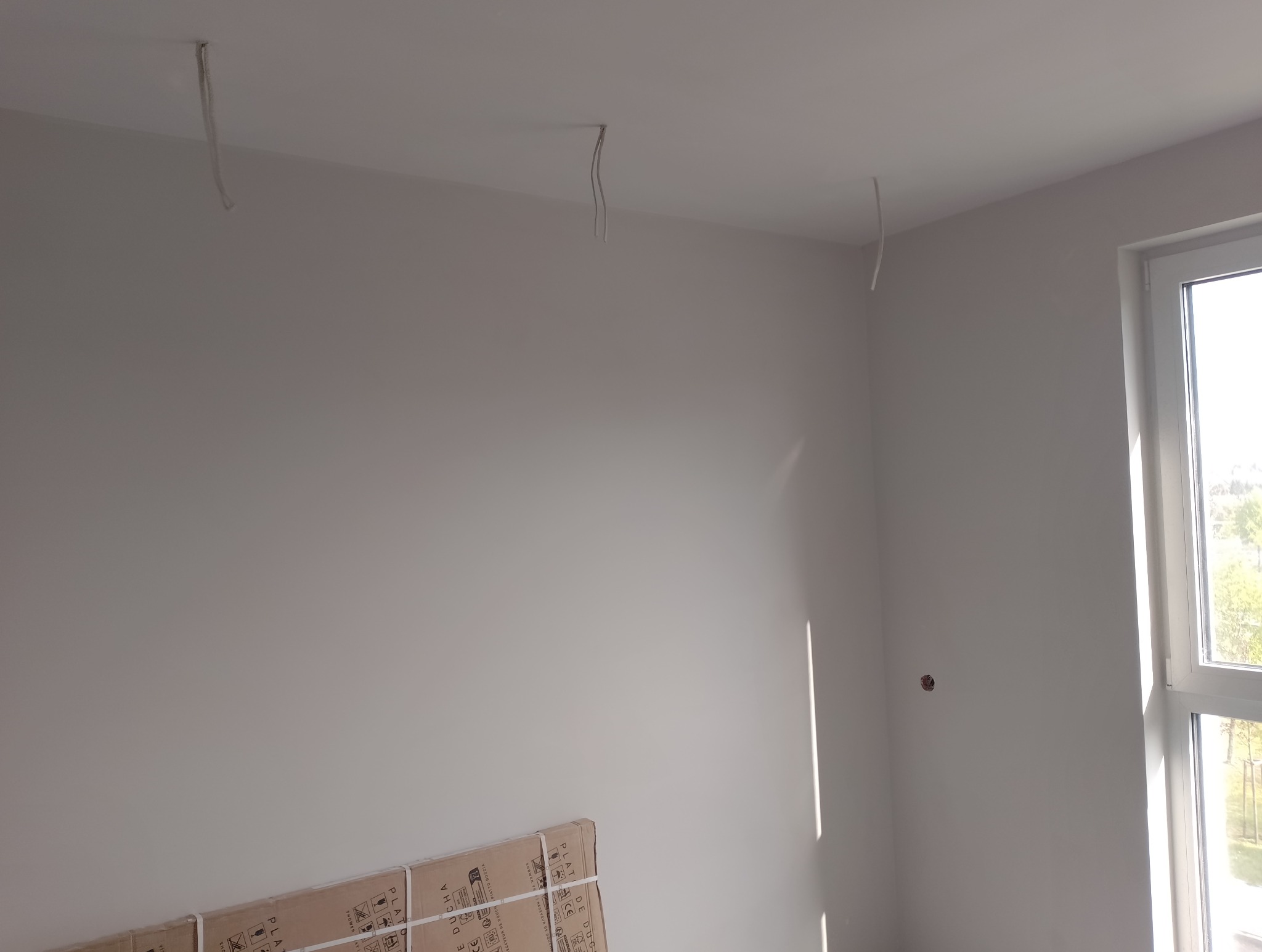 Pokój w trakcie prac wykończeniowych: gładkie, białe ściany, wystające kable z sufitu i kartony z elementami wyposażenia łazienki przy ścianie.