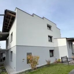 Dwukolorowa elewacja budynku po remoncie: górna część biała, dolna szara, z widocznymi oknami w brązowych ramach i fragmentem ogrodu z niskim, metalowym ogrodzeniem.