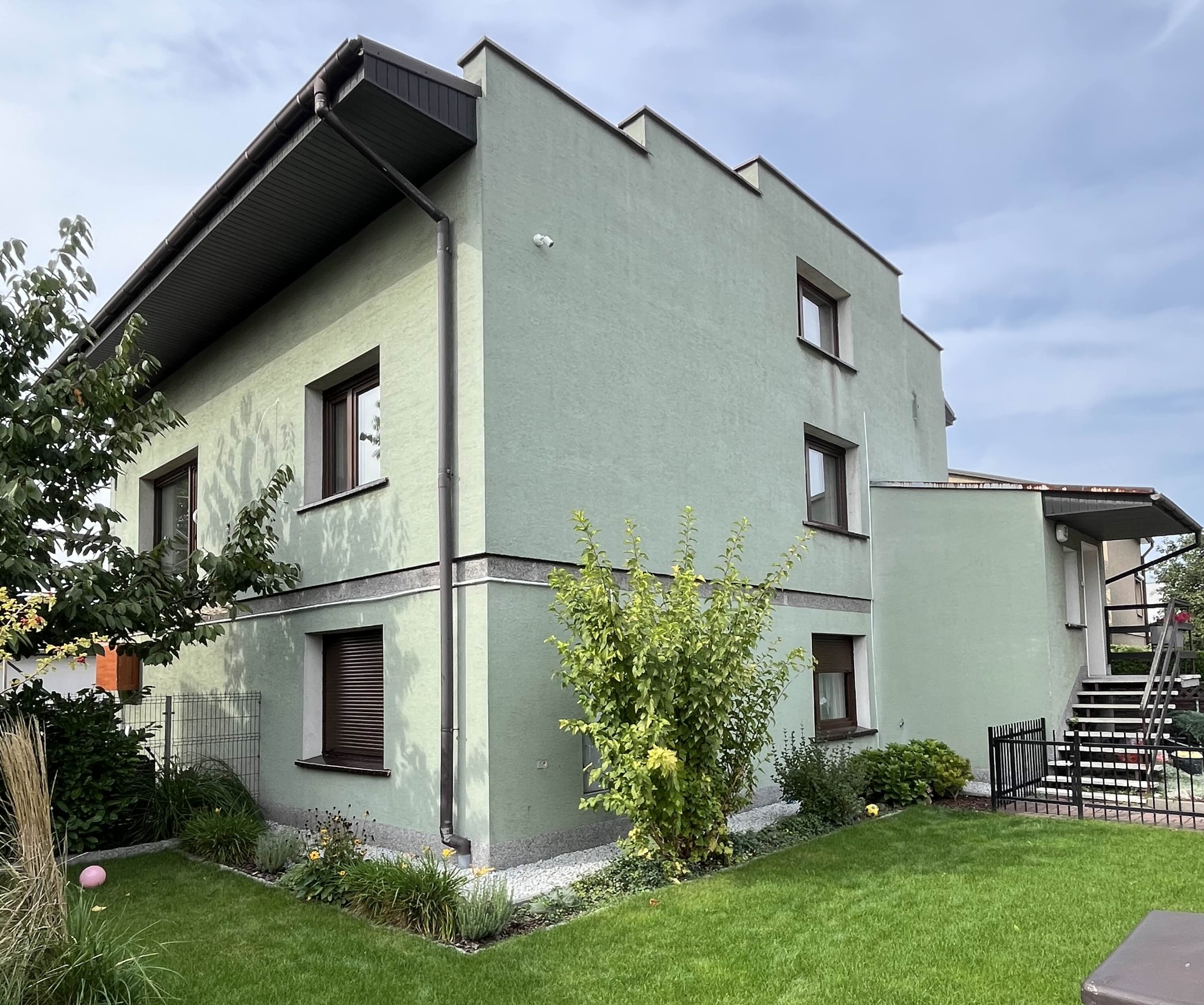 Dwupiętrowy dom z elewacją w odcieniach zieleni, brązowe okna z roletami, zadbany trawnik i ozdobne krzewy w ogrodzie.