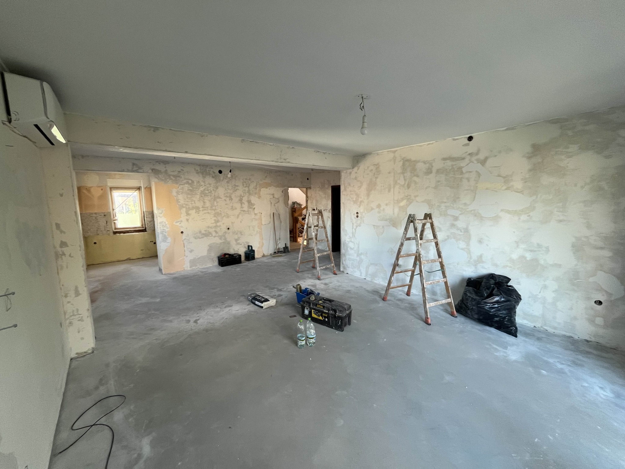 Wnętrze w trakcie remontu: przygotowane ściany z widocznymi śladami szpachlowania, dwie drabiny, torba z odpadami i skrzynka z narzędziami na betonowej podłodze. W tle widoczne okno i dalsze...