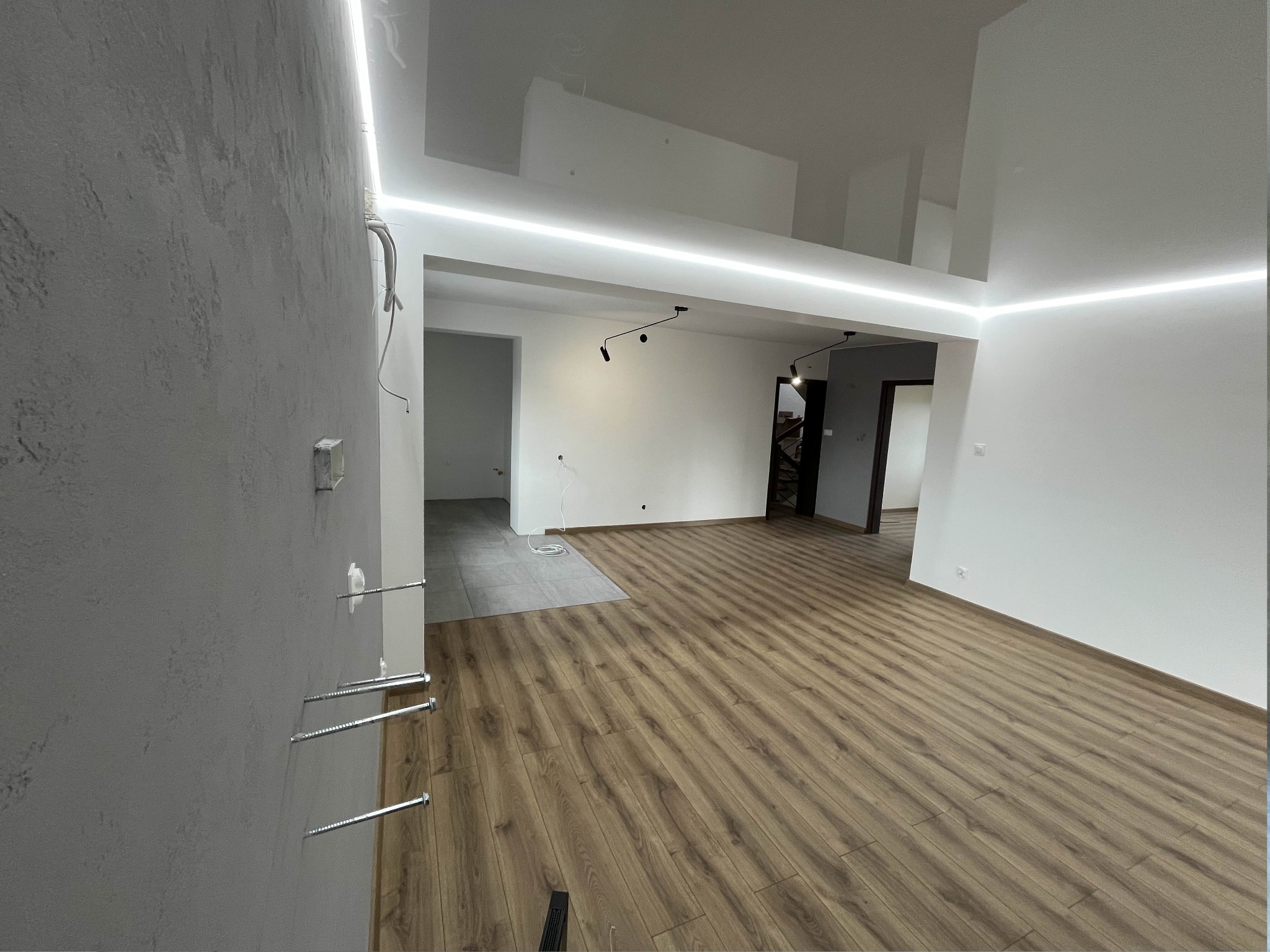 Wnętrze mieszkania w trakcie prac: wykończona podłoga z paneli, surowa ściana z widocznymi kołkami montażowymi, oświetlenie LED podsufitowe, fragment położonych płytek w przejściu do innego...