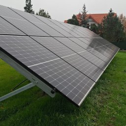 E&R - EvaRaf - Rafał Adamczyk - Rząd paneli słonecznych na metalowej konstrukcji, zainstalowany na trawniku przed domem z czerwoną dachówką w pochmurny dzień.