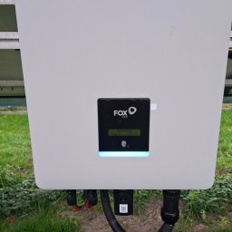 E&R - EvaRaf - Rafał Adamczyk - Biała obudowa inwertera Fox ESS z wyświetlaczem wskazującym moc 252W, zamontowana na tle zielonej trawy, widoczne kable i złącza.