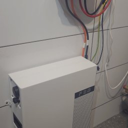 E&R - EvaRaf - Rafał Adamczyk - Biała szafka magazynu energii FEB z podłączonymi kablami elektrycznymi i zabezpieczeniem Noark na ścianie z poziomymi panelami.