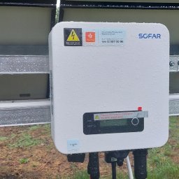 E&R - EvaRaf - Rafał Adamczyk - Biały inwerter fotowoltaiczny marki Sofar, mokry od deszczu, zamontowany na metalowej konstrukcji obok paneli słonecznych, widoczne naklejki ostrzegawcze i informacyjne.