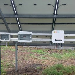 E&R - EvaRaf - Rafał Adamczyk - Widok od spodu na zamontowane panele słoneczne z widocznymi puszkami przyłączeniowymi i inwerterem Sofar, zainstalowane na metalowej konstrukcji na zewnątrz w pochmurny dzień.