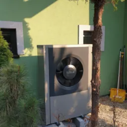 Szara pompa ciepła Buderus na betonowych podstawach, ustawiona przed zieloną ścianą domu z dwoma oknami, częściowo zasłonięta przez iglaste krzewy i małe drzewko.